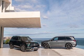Two Mercedes GLS models in deep forest green and gray, photographed next to a modern house with the ocean behind them.