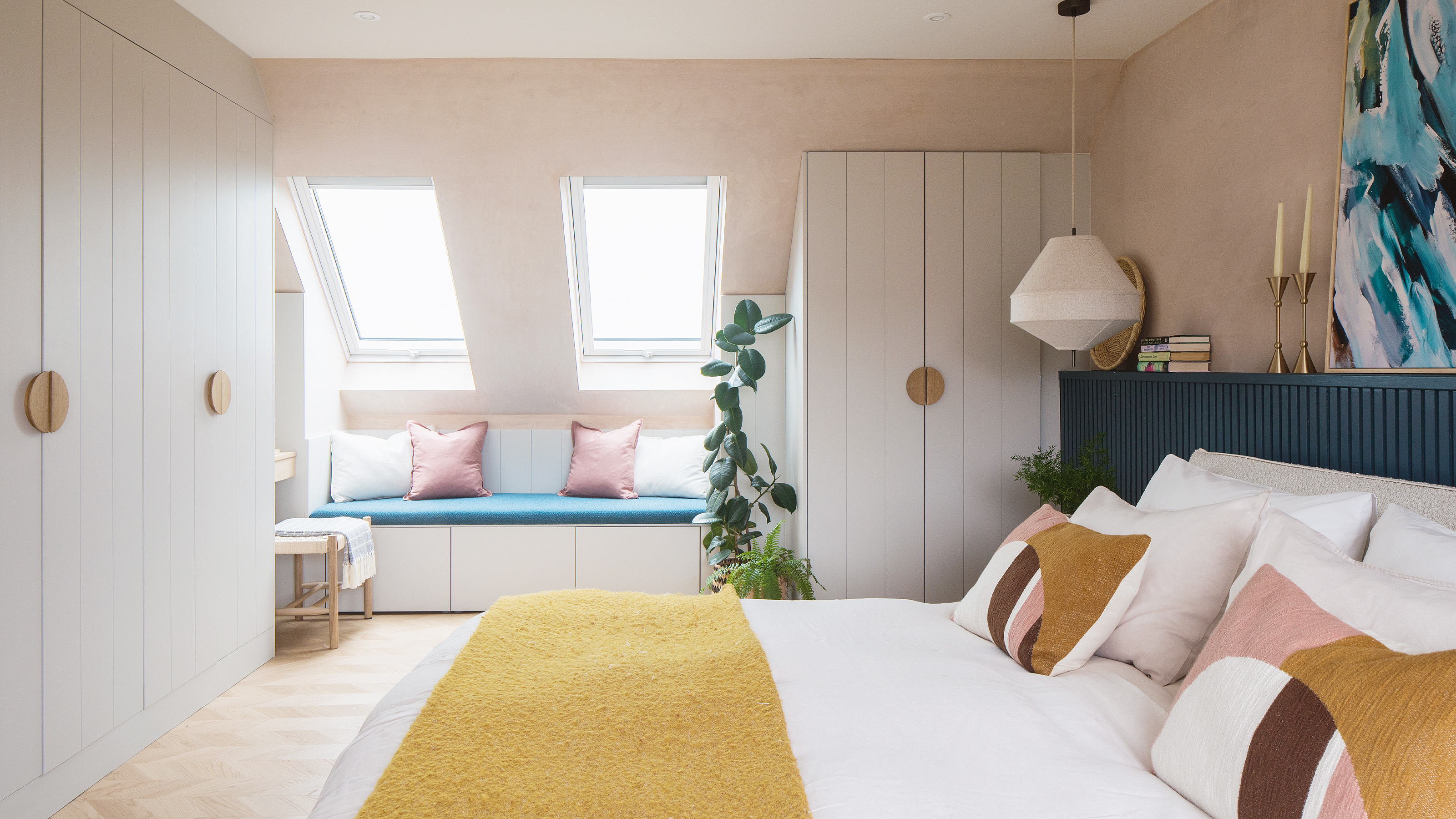 A colourful bedroom with dormer windows and pale pink walls. The double bed is dressed in white bedding with yellow, pink and burgundy accents.