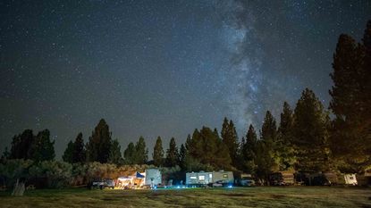 a collection of recreatonal vehicles and tents on a grassy field surrounded by pine trees at night beneath a starry sky and a vertical milky way