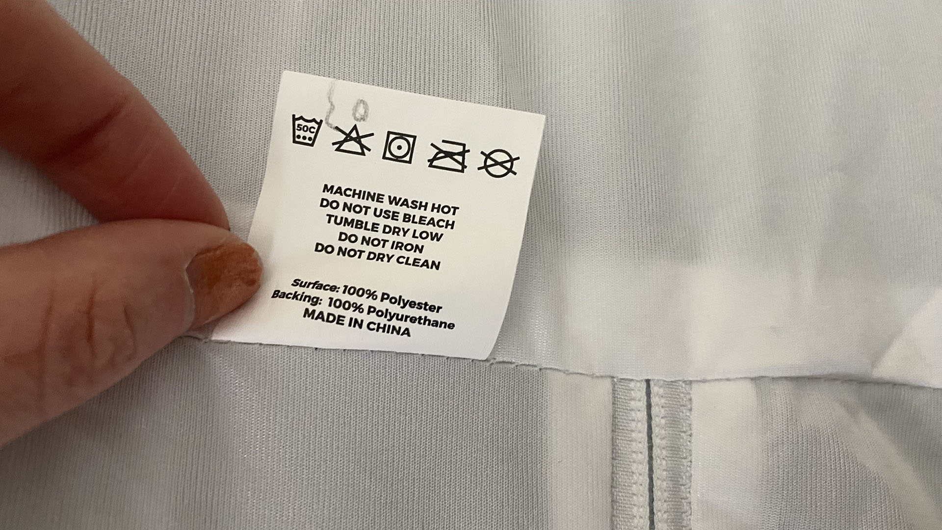 A closeup of fingers holding the Linenspa Mattress Encasement care instructions tag, which read: machine wash hot, do not use bleach, tumble dry low, do not iron, do not dry clean