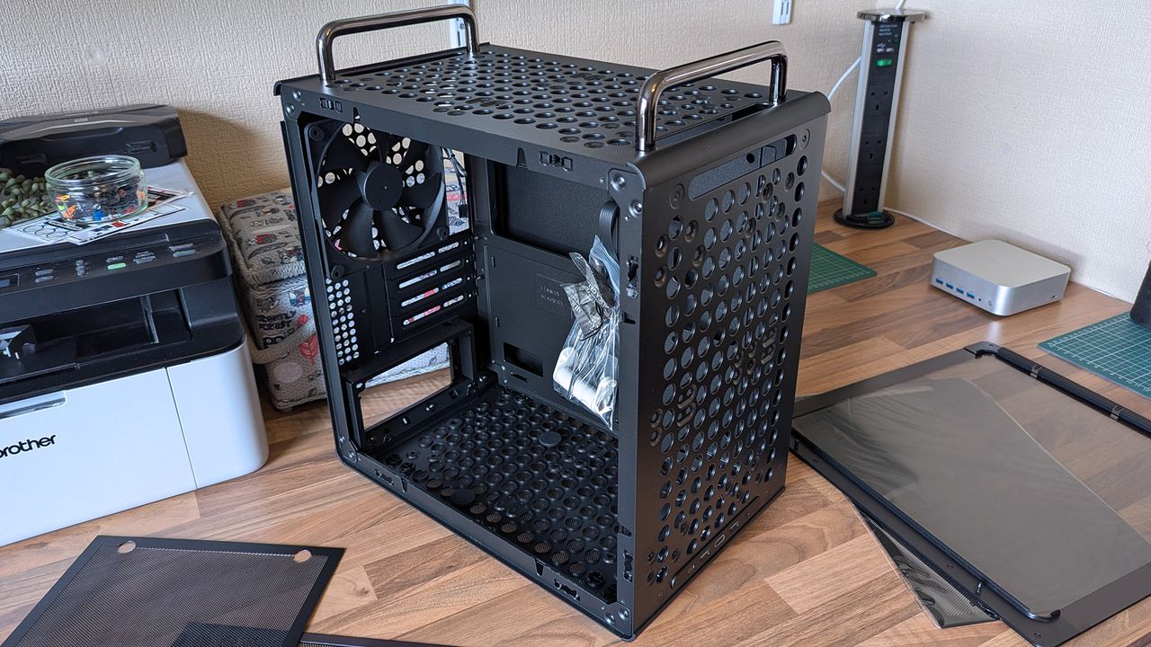 A Cooler Master Qube 340 case sits on a desk with its side panels removed, showing the empty interior, rear fan, and a small bag of screws inside the chassis.