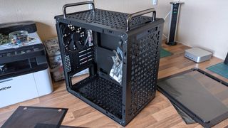 A Cooler Master Qube 340 case sits on a desk with its side panels removed, showing the empty interior, rear fan, and a small bag of screws inside the chassis.