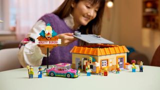 A woman smiles as she lifts the roof from the Lego Krusty Burger set, which sits on a plain surface