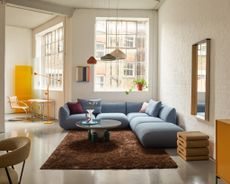 A naturally lit, lofty design showroom is filled with shapely late 20th-century furniture, including a light blue sofa, a fluffy brown rug, a wavy bedside table, and yellow cabinets.