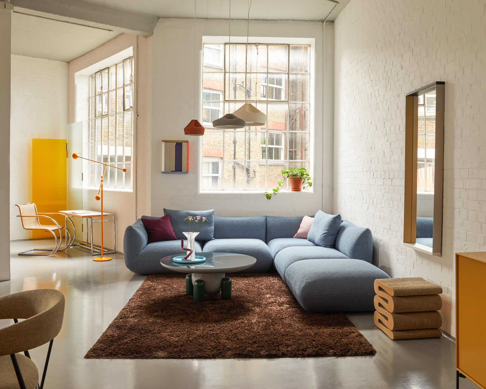 A naturally lit, lofty design showroom is filled with shapely late 20th-century furniture, including a light blue sofa, a fluffy brown rug, a wavy bedside table, and yellow cabinets.