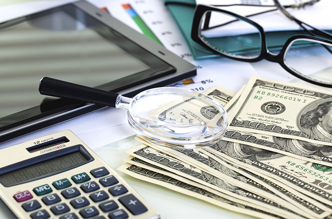 Pile of cash, a laptop, eyeglasses and paperwork on a desktop