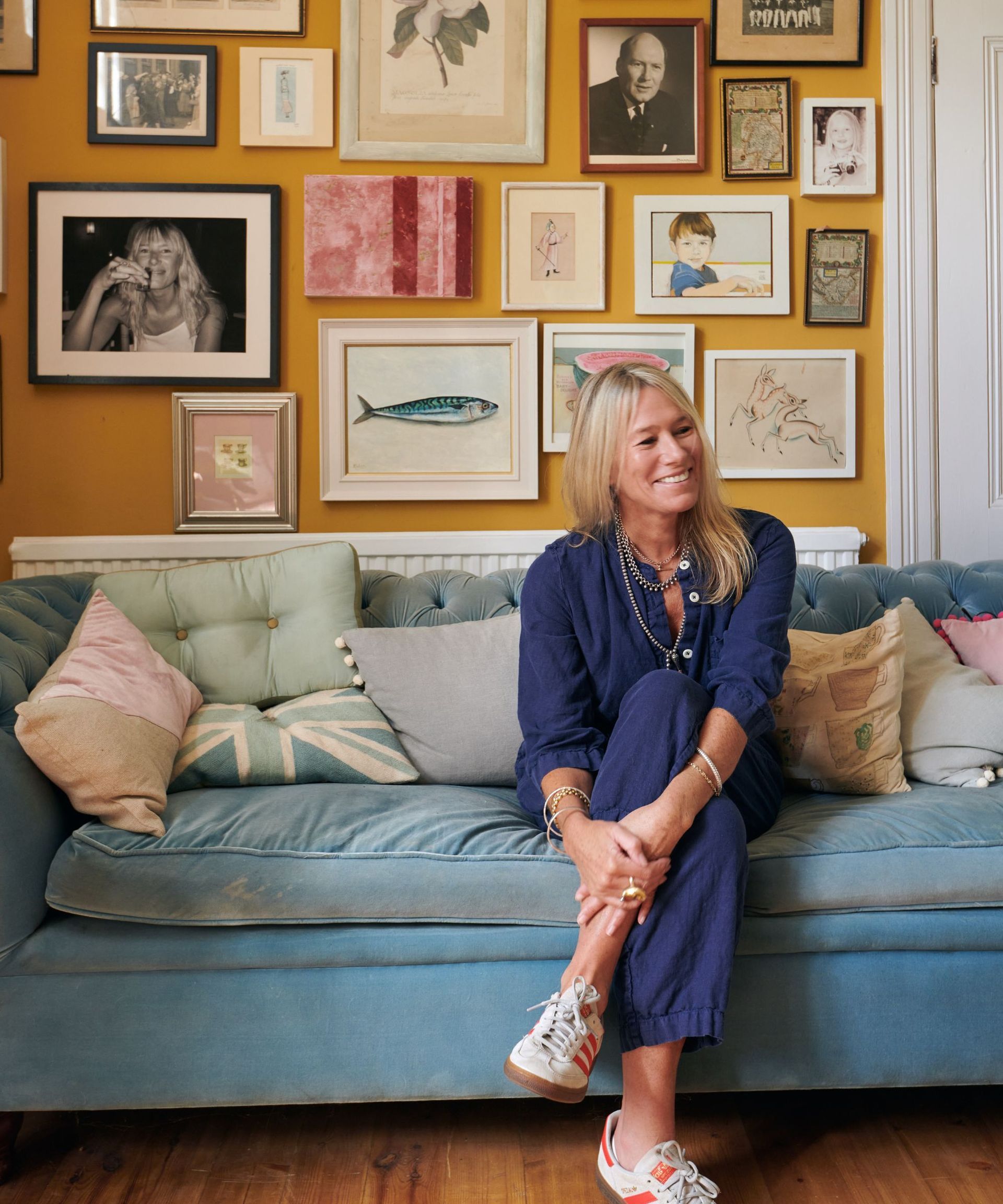 A blonde woman in a navy boiler suit sits on a baby blue velvet sofa with a picture gallery wall behind
