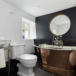 Cotswold cottage's bathroom with copper bath, large round mirror and black feature wall