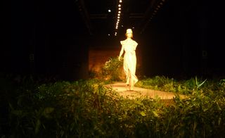 Model wearing a dress and hat, on a runway lined with foliage