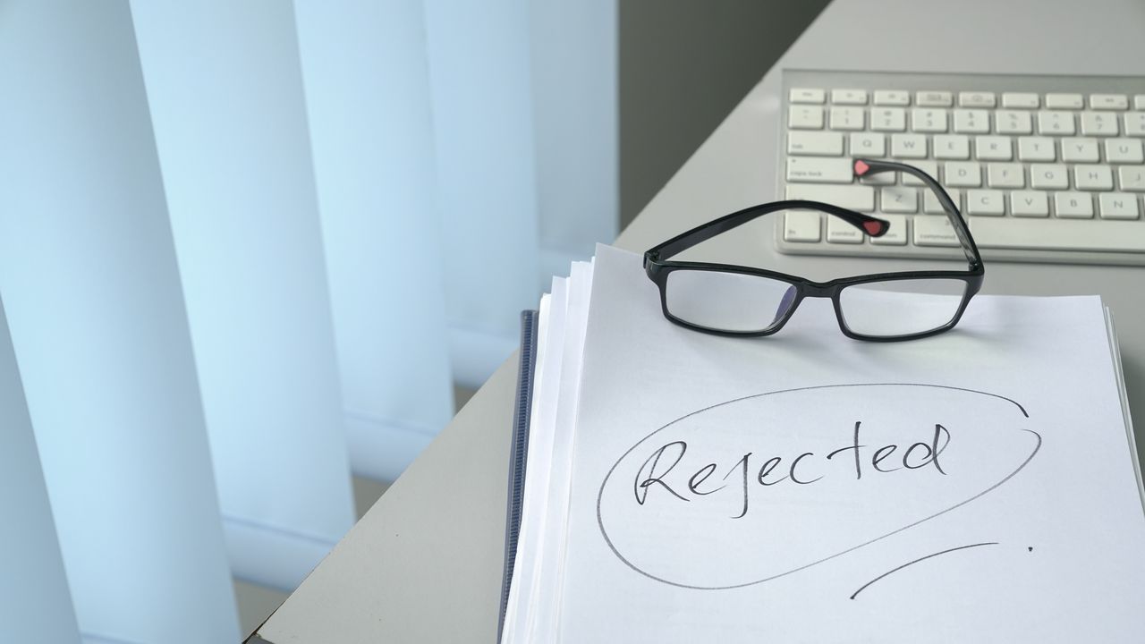 On a desk next to a computer keyboard, eyeglasses sit on a stack of papers. The word &quot;rejected&quot; is written on the top page.