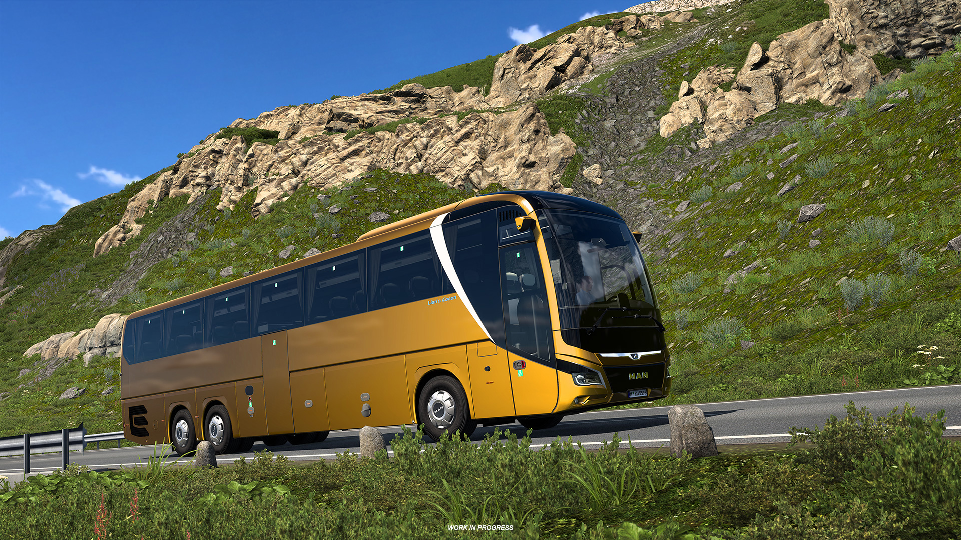 A gold MAN Lion&rsquo;s Coach drives along a winding mountain road in Euro Truck Simulator 2, surrounded by rocky hills and greenery under a bright blue sky, with a small &ldquo;Work in Progress&rdquo; label on screen.