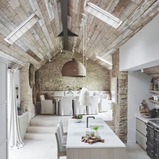 white rustic kitchen with exposed brick walls, reclaimed wood ceiling, white marble top island, large wicker pendant light, covered seats, large dining table, bench seating at rear