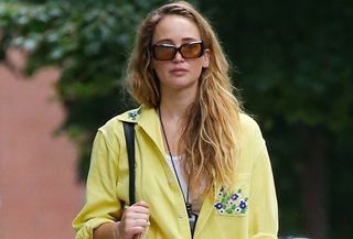 Jennifer Lawrence wearing a yellow Bode button-down shirt with sunglasses, a white tank top, white trousers, a Loewe raffia tote, and Melissa jelly shoes.