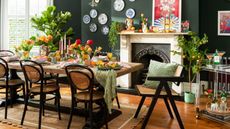 Narrow dining room ideas can be stylish, too. Here is a dark green dining room with a long wooden table with colorful candles, flowers, and tableware on it, with two wooden and black iron curved chairs underneath it and a scalloped jute rug underneath that