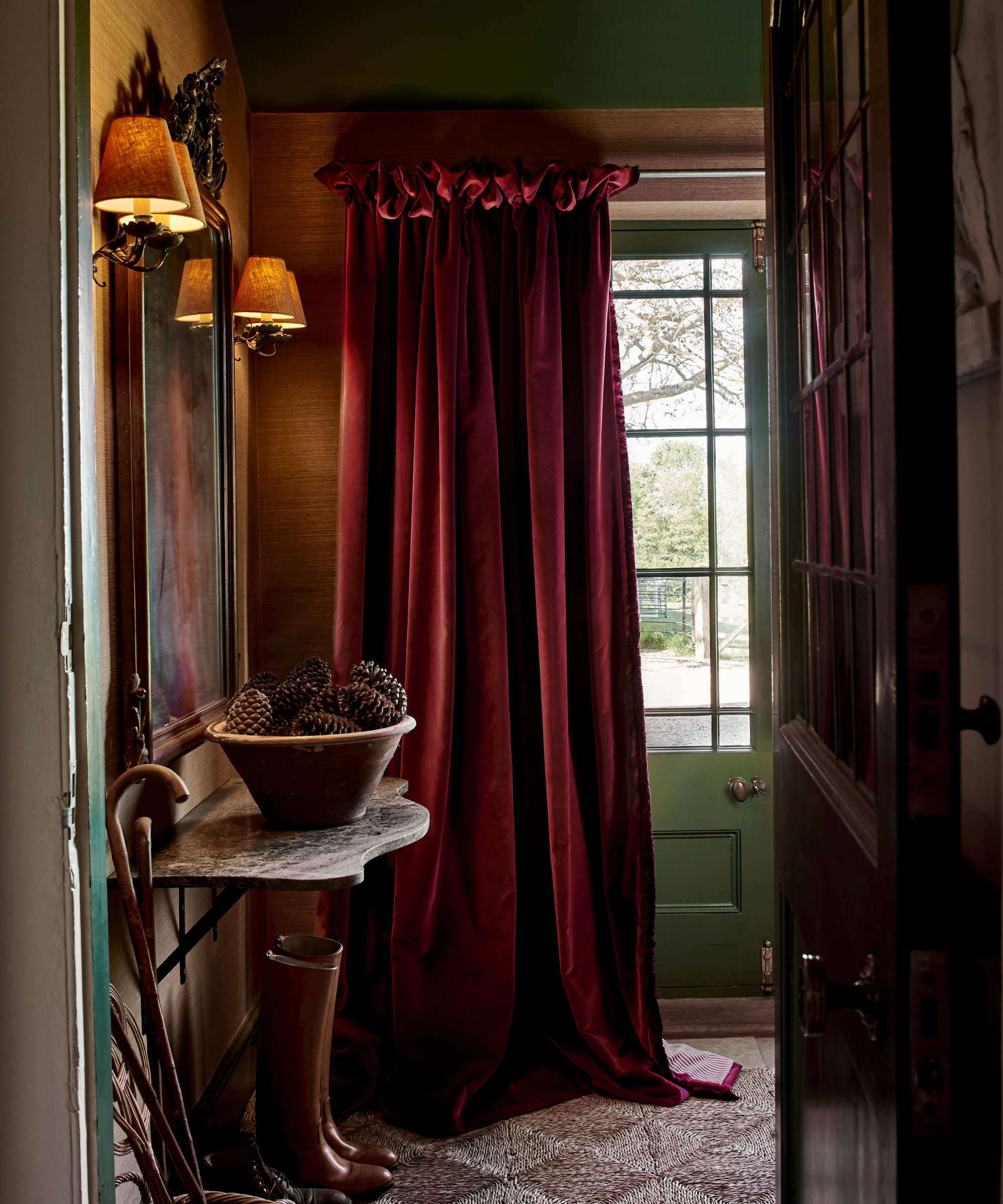 an entryway with a dramatic red velvet curtain, a marble table holding a bowl of pinecones, an artwork hanging against the wall with two sconces on either side, and a pair of boots on the floor