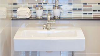 Rectangular white wall hung sink with chrome tap and glass shelf with accessories against a small rectangular tiles splashback