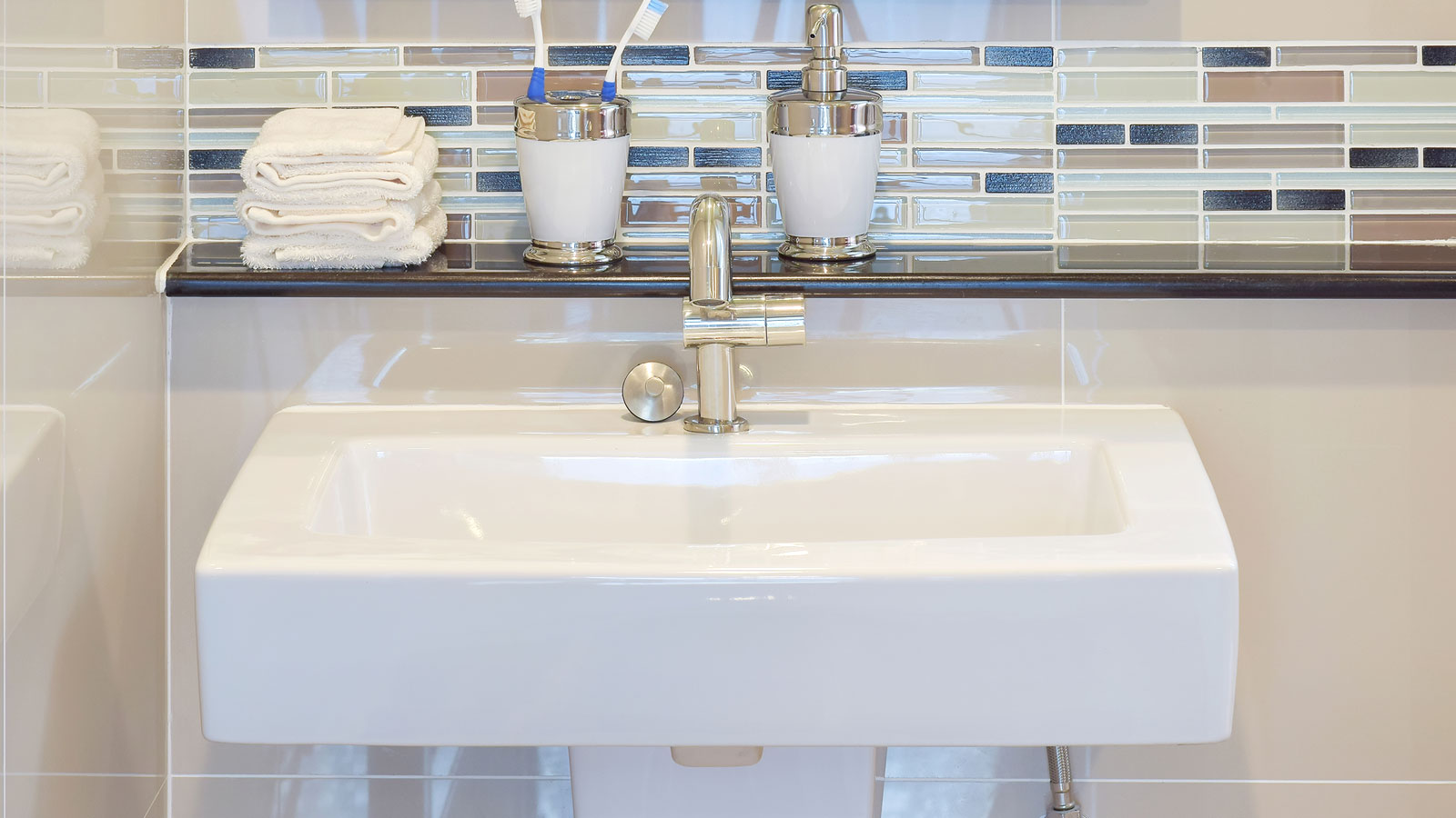 Rectangular white wall hung sink with chrome tap and glass shelf with accessories against a small rectangular tiles splashback