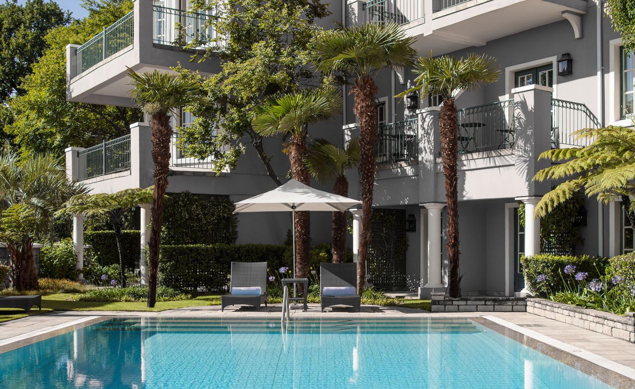 Swimming pool area at the Four Seasons Hotel. Two loungers with a sun umbrella is facing the pool and it is surrounded by palm trees and a beautiful garden.