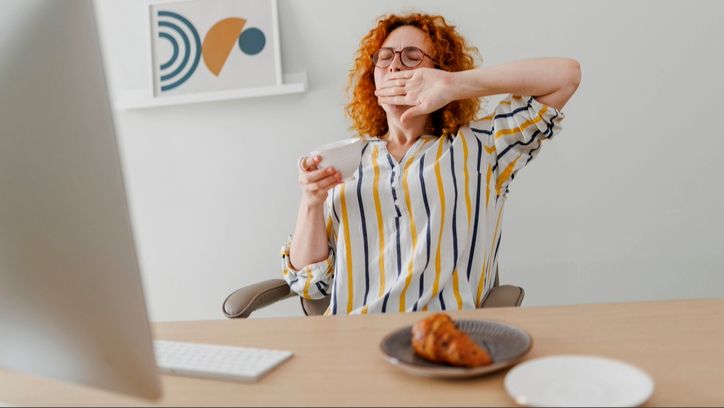 A woman with red hair wearing a white, yellow and navy stripey shirt sitting at desk yawning with a mug in hand and a pastry on plate on desk in front of computer