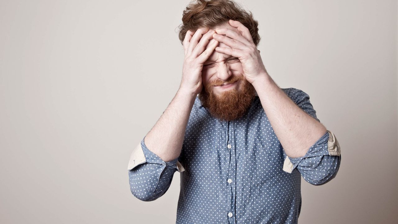 A young man with a beard holds his head in his hands in dismay.