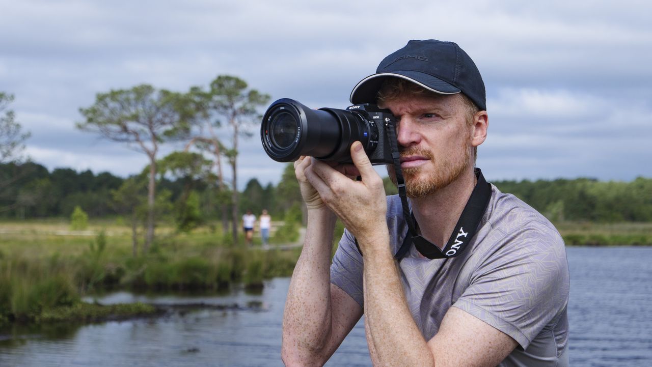 Photographer and tech journalist Tim Coleman holding the Sony RX10 V bridge camera up to his eye in a nature reserve, with dramatic clouds behind him