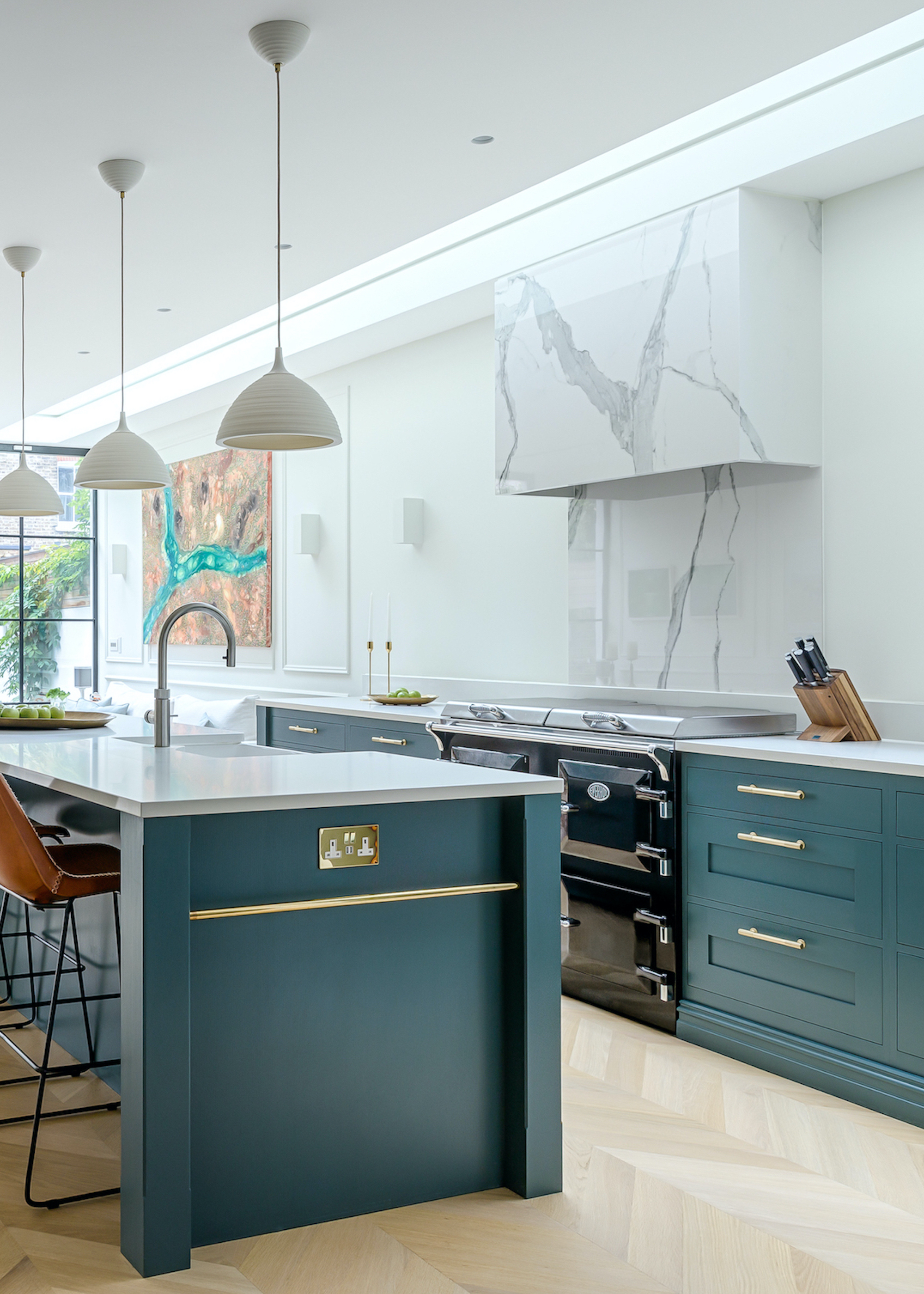 A blue kitchen island with gold features and blue kitchen cabinets with gold handles. The kitchen island has a sink and above it are three pendant lights.