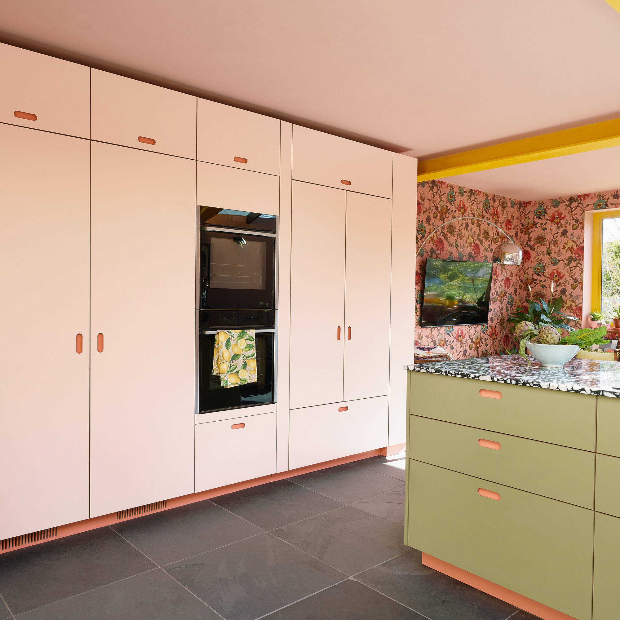 kitchen with wall of full height pale pink cabinets with integrated double oven grey tiles flooring and pensinula island wiht green cabinetry and black and white worktopscolourful kitchen with side extension pink ceiling yellow steel beams yellow framed window and glass door floral pink wallpaper and green kitchen cabinets with black and white worktop