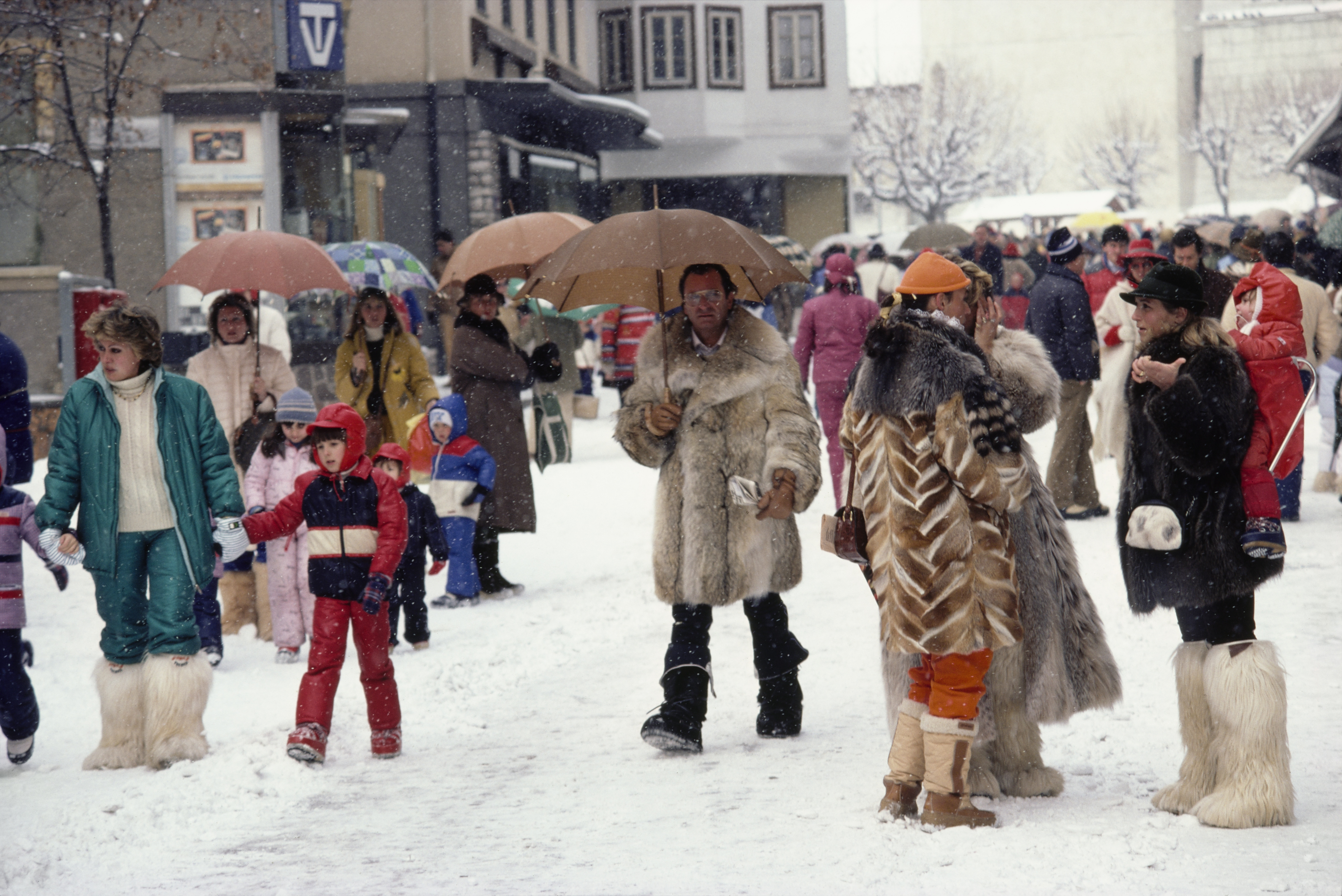 A busy snowy village street scene, with people in winter clothing, umbrellas raised, and several figures wearing fur-trimmed coats and boots.