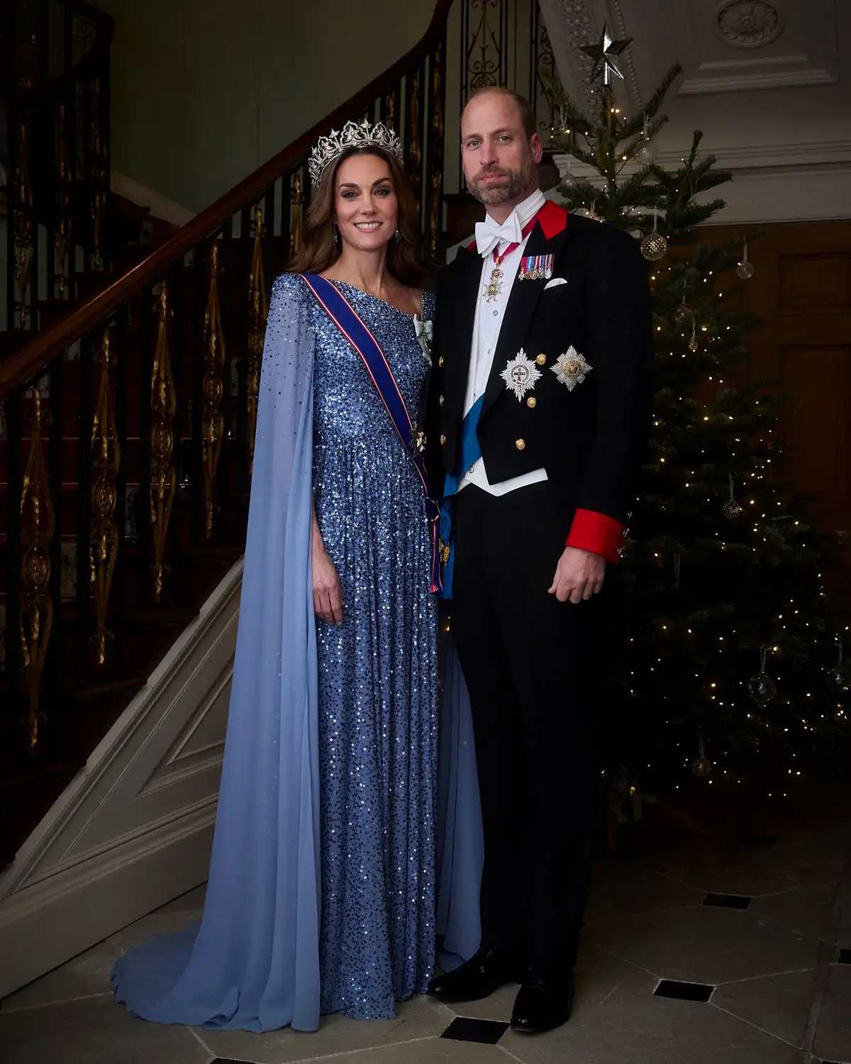 Kate Middleton and Prince William standing in front of a Christmas tree in a blue sequin gown and suit