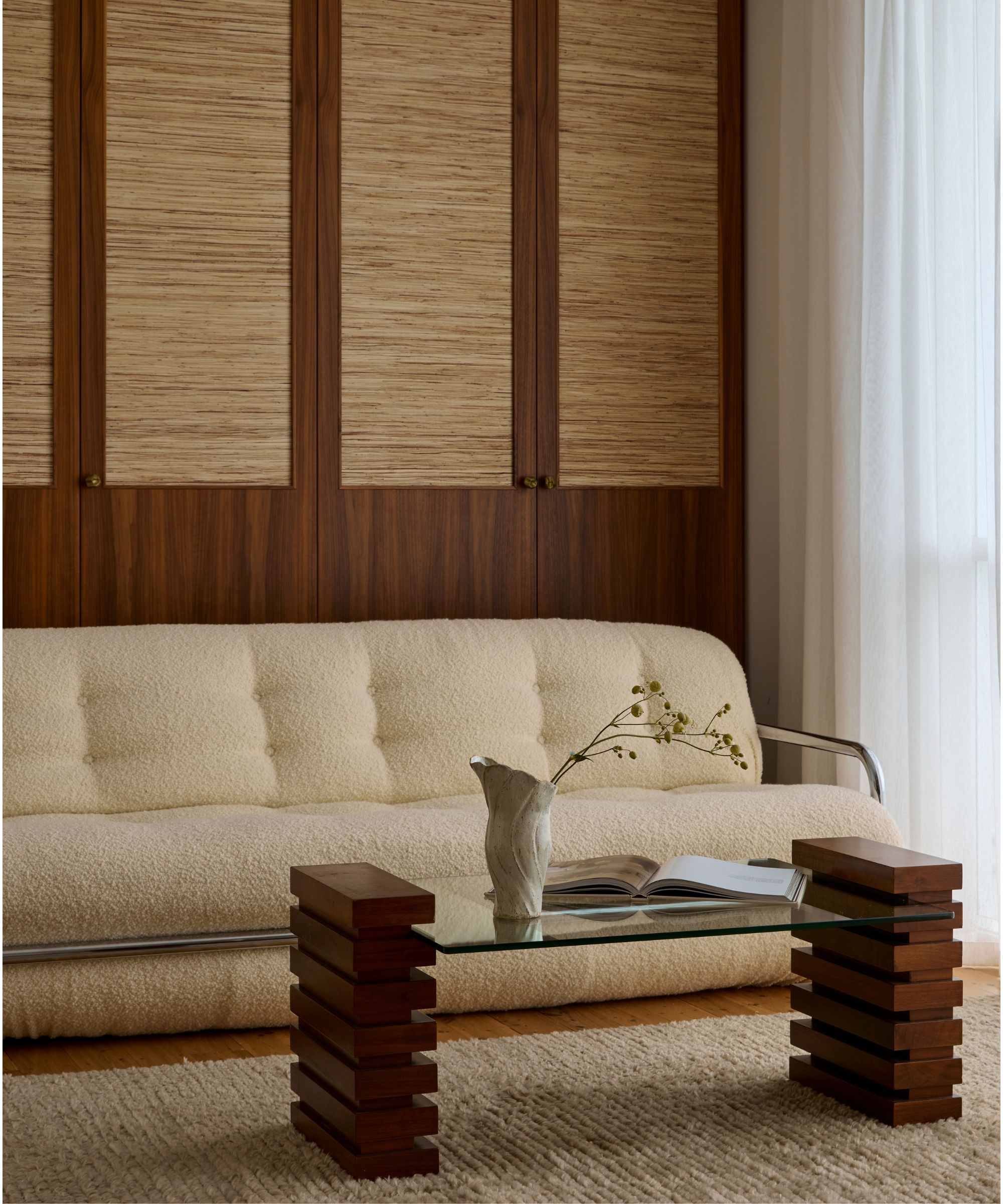 1970s Italian boucl&amp;eacute; sofa with chrome frame, styled with a glass coffee table on stacked wooden supports, set against woven-paneled cabinetry and sheer curtains.
