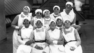 A group of ten nurses wearing their uniform and round dark glasses all smile and look up to the sky. 