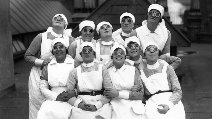 A group of ten nurses wearing their uniform and round dark glasses all smile and look up to the sky. 