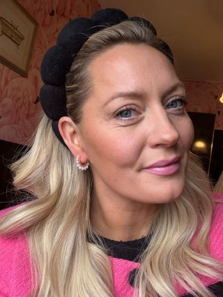 A headshot of Sophie McGown in a wallpapered room wearing a pink coat and black headband smiling and showing off a pair of Catherine beaded hoop earrings