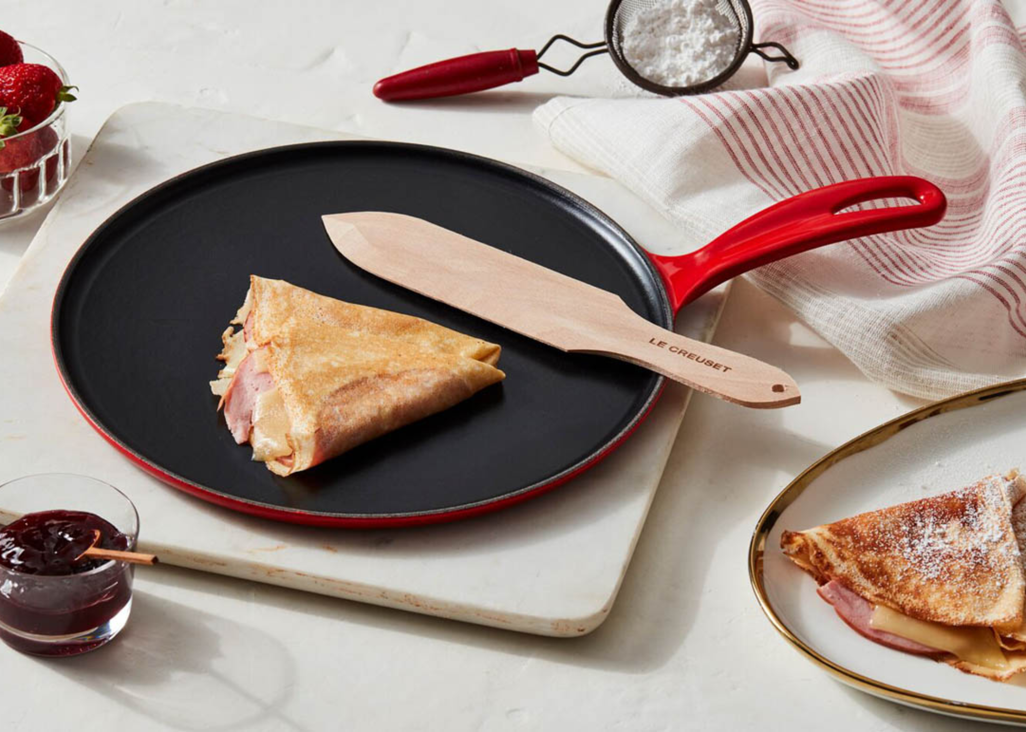 A red cast iron crepe pan on a kitchen board with a crepe and a plate of crepes