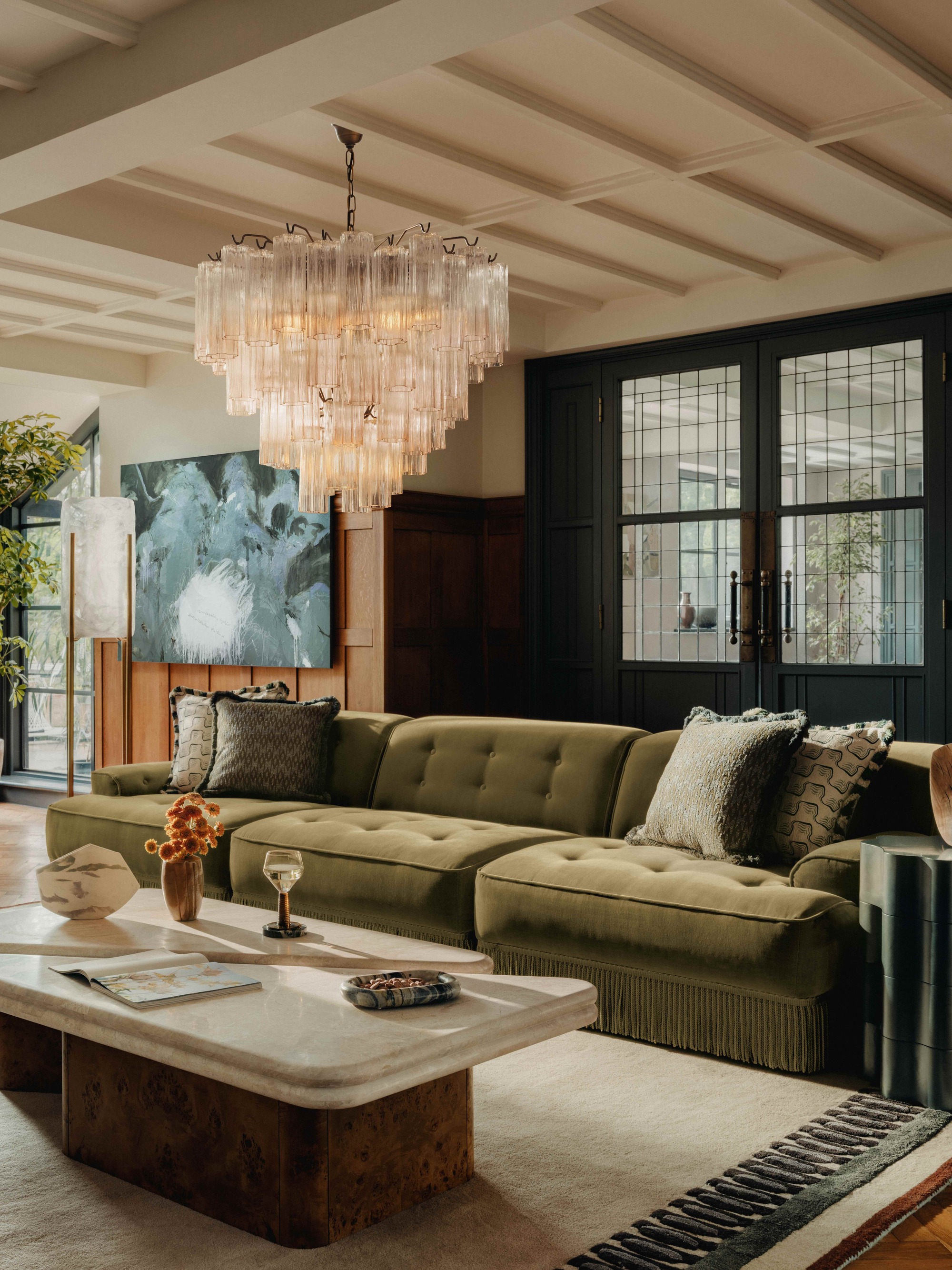 modern living room with decorative ceiling details, chandelier, white rug with black pattern, stone coffee table, green velvet sofa with fringe detailing