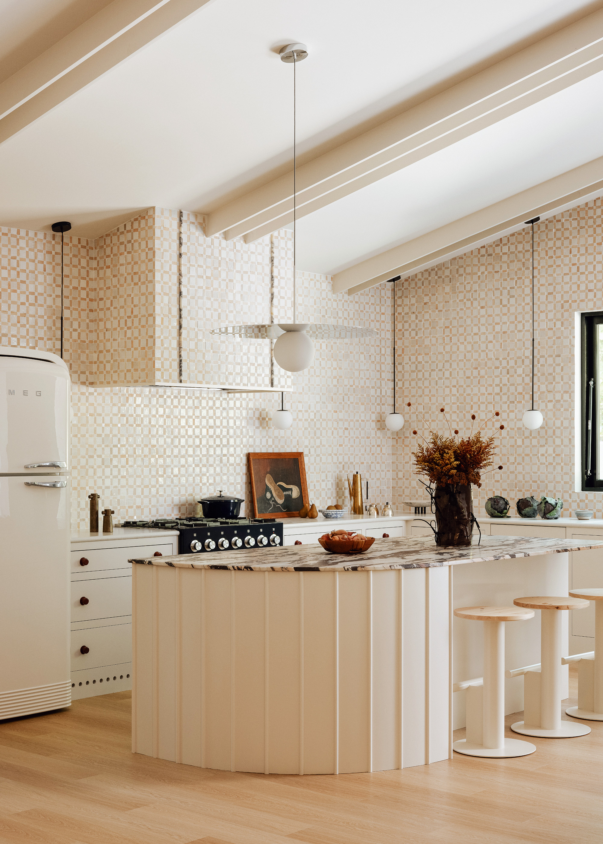Off-white kitchen with glossy cream zellige tiled walls, curved island with black and white marble countertop, and cabinetry with burgundy hardware.