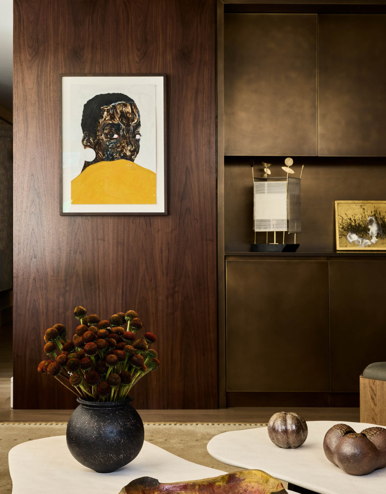 A living room with a dark wood wall and a framed art piece by bronze cabinets and ahead of a coffee table with a vase of flowers
