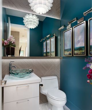 A teal blue and white bathroom with a white sink base with a glass wavy sink, a white toilet, green and blue countryside prints on the righthand wall, a large mirror on the center mirror, and a white sculptural light