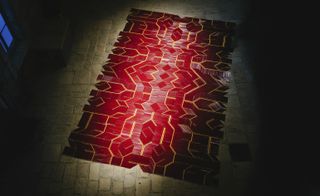 Dimly lit interior of Hansen House’s courtyard, stone brick floor, carpet rug made from 12,000 red and yellow pencils with spotlight shining over it