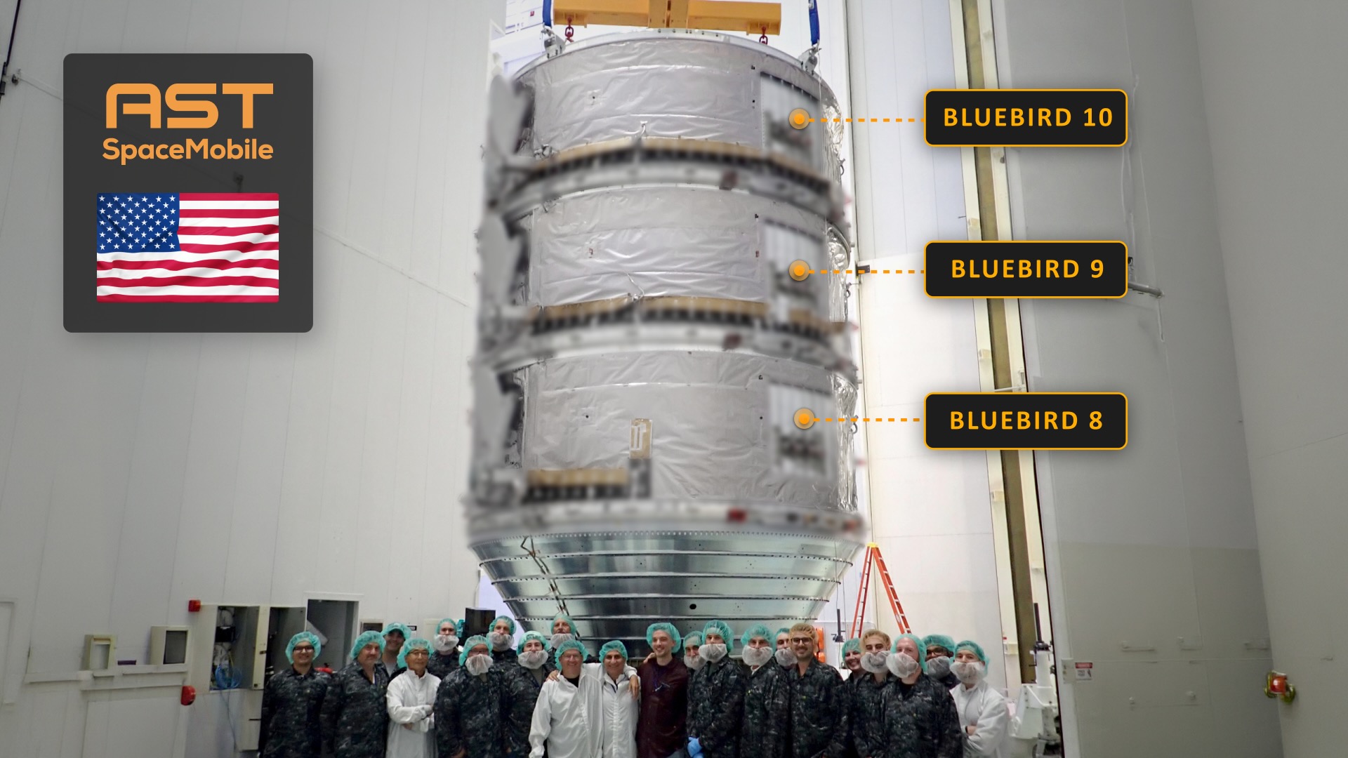 Em uma grande sala com paredes brancas, dezenas de pessoas estão diante de uma pilha de três grandes satélites