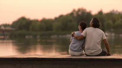 A retired couple sit on the side of a dock on a lake and watch the sun set.