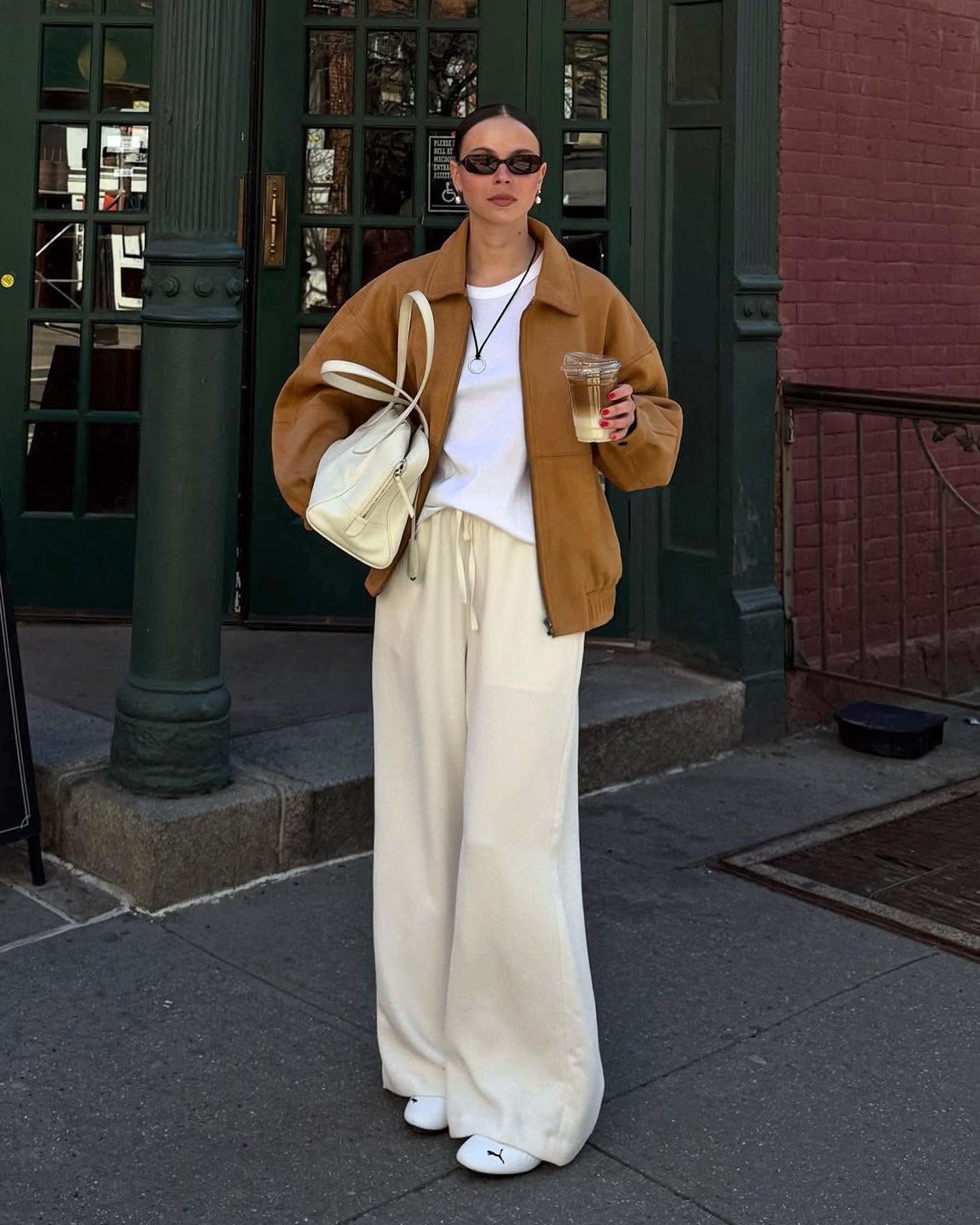 Scandi style influencer Johanna Lager poses in the West Village of NYC wearing oval sunglasses, an oversize tan suede bomber jacket, a black cord pendatn necklace, oversize white T-shirt, cream bowler bag, ivory satin drawstring pants, and Puma Speedcat sneakers