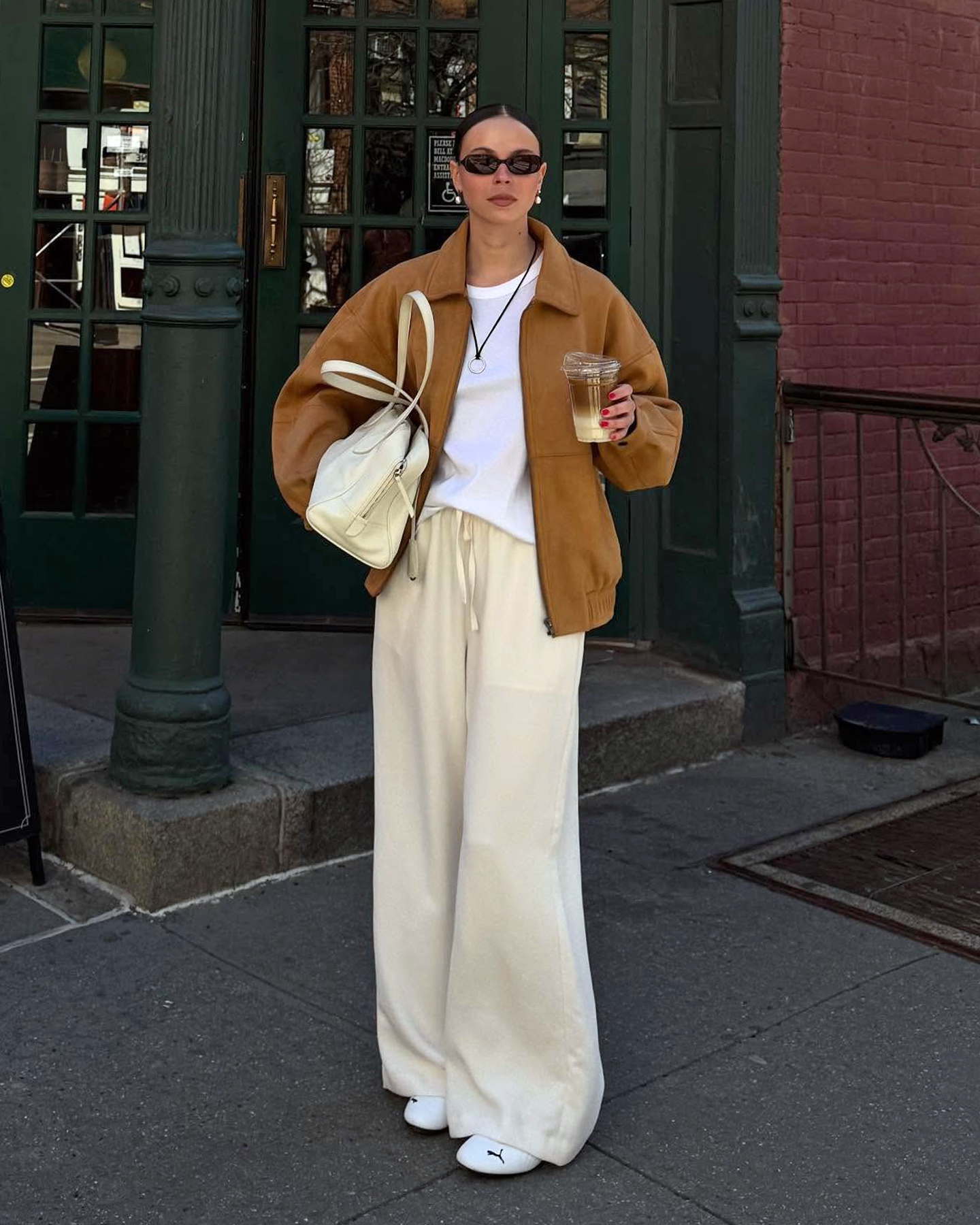 Scandi style influencer Johanna Lager poses in the West Village of NYC wearing oval sunglasses, an oversize tan suede bomber jacket, a black cord pendatn necklace, oversize white T-shirt, cream bowler bag, ivory satin drawstring pants, and Puma Speedcat sneakers