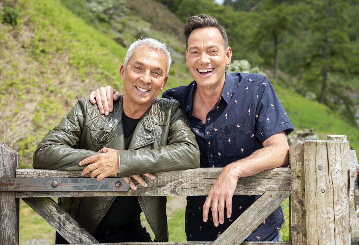 Bruno Tonioli and Craig Revel Horwood pose for a photo while leaning on a fence gate in a lush green landscape on a sunny day 