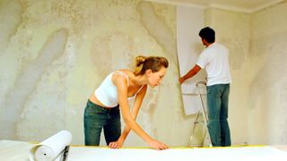 Two people, one hanging wallpaper on a bare wall, one using a pasting table 
