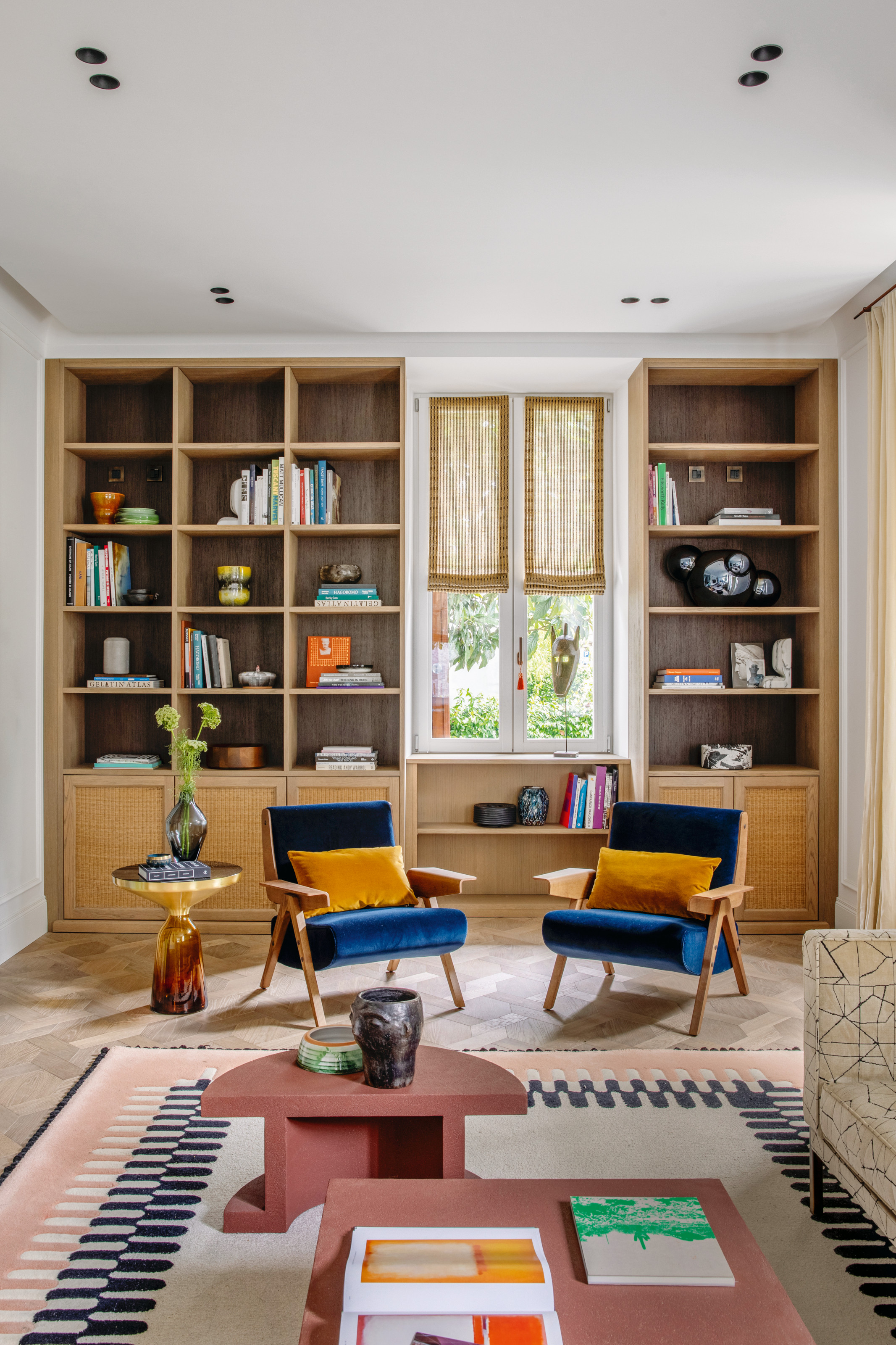 Living room with built-in wooden shelving, terracotta two-piece geometric coffee table, electric blue armchairs and white, black and pale pink patterned rug