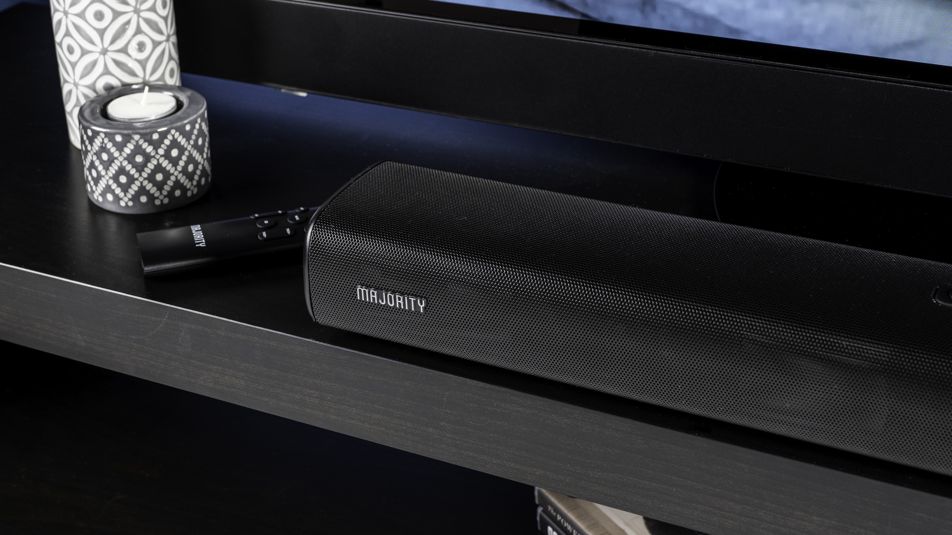 majority elias, a black soundbar with a silver majority logo, and a hdmi earc port and optical port, with a black remote, photographed on a black TV stand