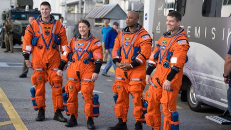 Four people in orange space suits stand on a road in front of a large van and look at a crowd off camera