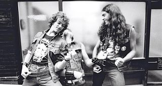 A black-and-white photo from the '80s of three heavy metal fans in full hesher garb, bullet belts, the lot, all playing air guitar in the street.