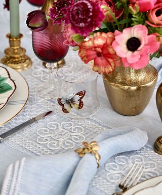 An outdoor dining table with a blue and white tablecloth, a pink floral arrangement and a glass decorated with a butterfly